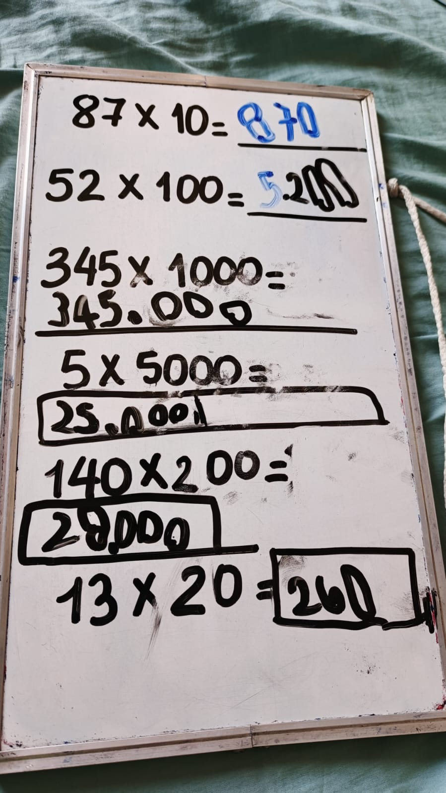 Pizarra acrílica con ejercicios de multiplicación por 10, 100, 1000 y múltiplos, resueltos a mano durante una sesión de matemáticas en casa